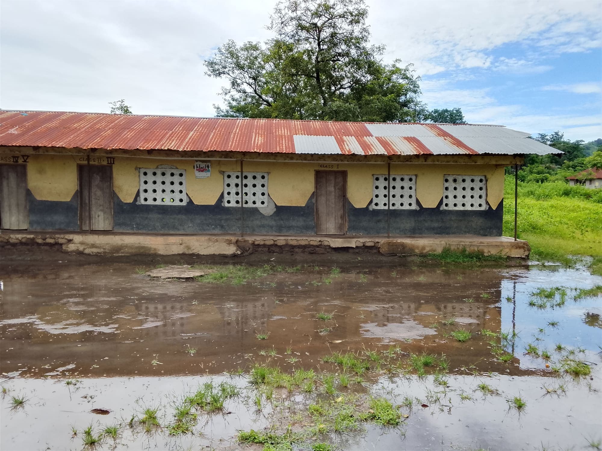 Side view of the school building with a few people standing on the veranda and wooden benches outside, on wet, muddy ground.