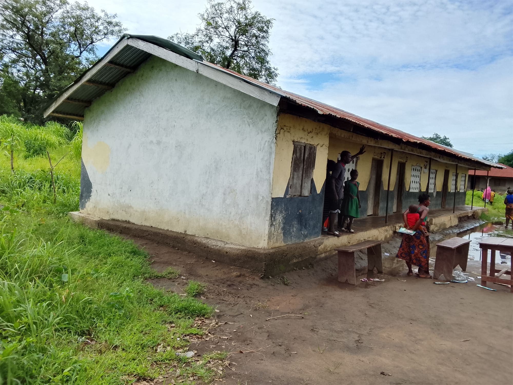 Front of a single-storey school building with a rusted corrugated roof and yellow walls. Standing rainwater covers the ground in front.