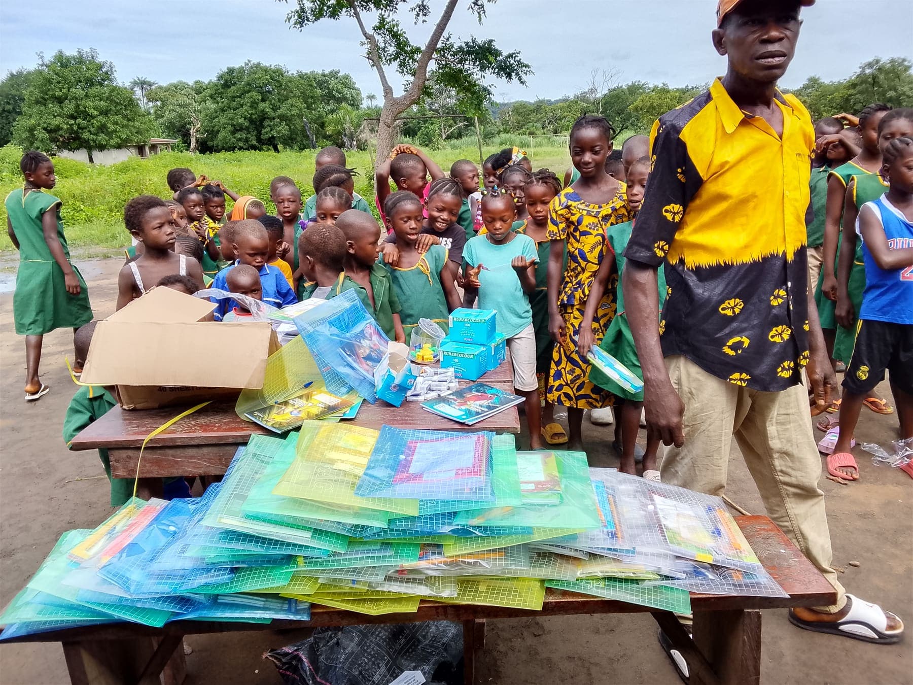 A man in a yellow shirt beside a table of school packs, with children in green school uniforms gathered behind.