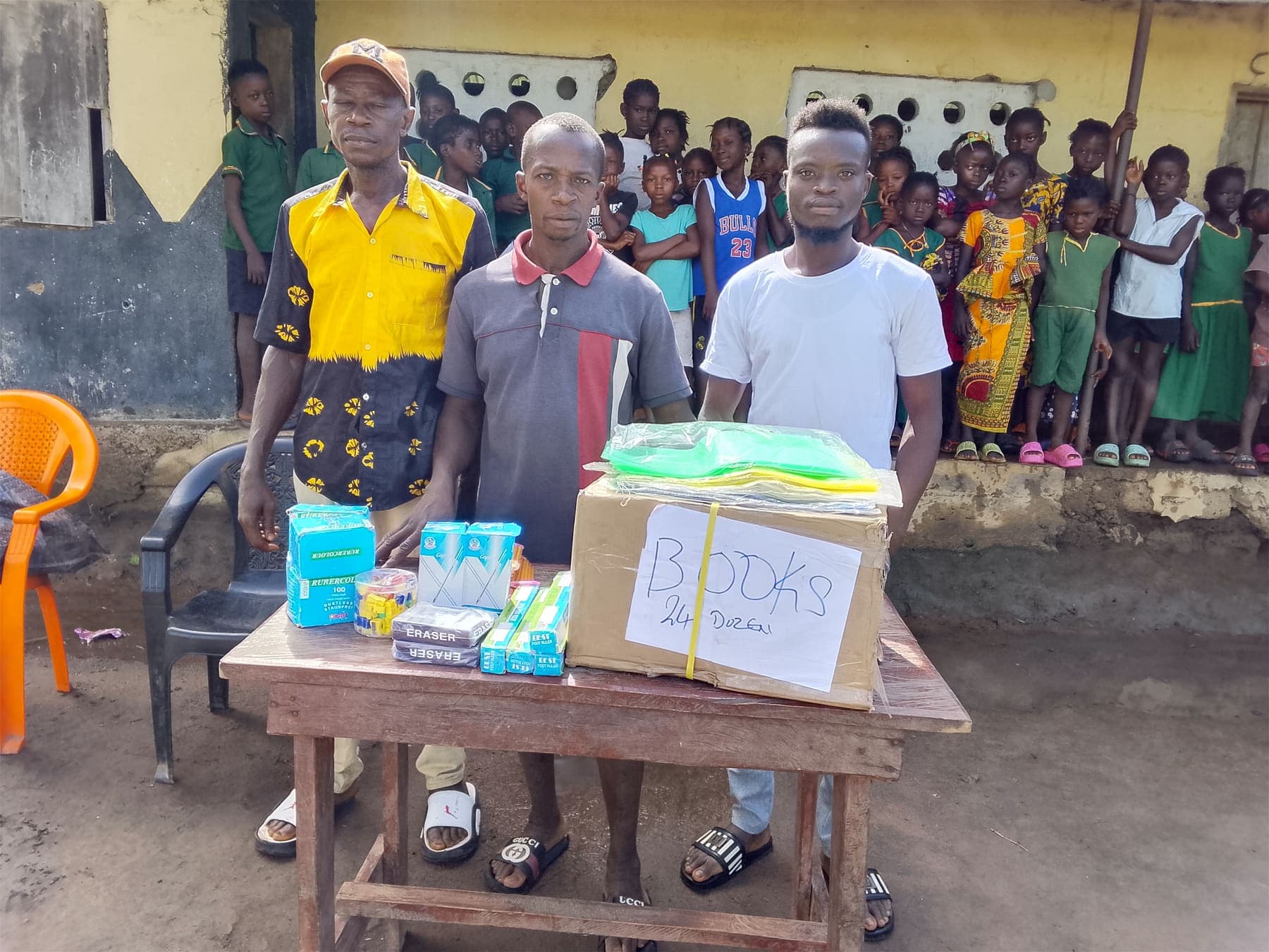 Three men standing behind a table with a box labelled books and school supplies, with pupils lined up behind them.