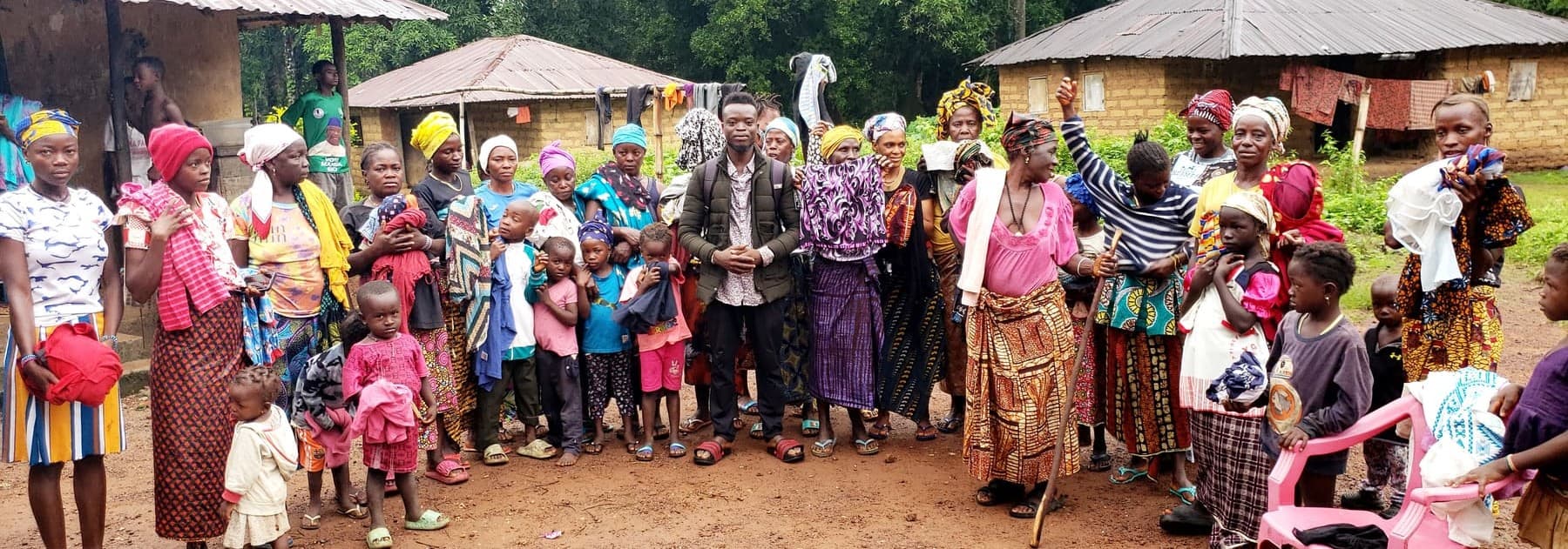 A large group of women and children in a village compound in Sierra Leone, many holding up items of clothing, with a CDEO team member standing in the middle.