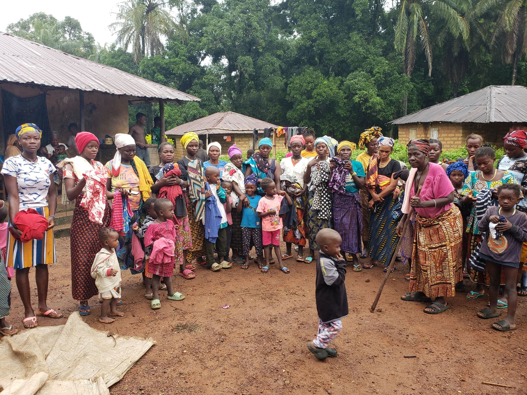 A large group of women, men, and children standing together in a village compound beside thatched houses.