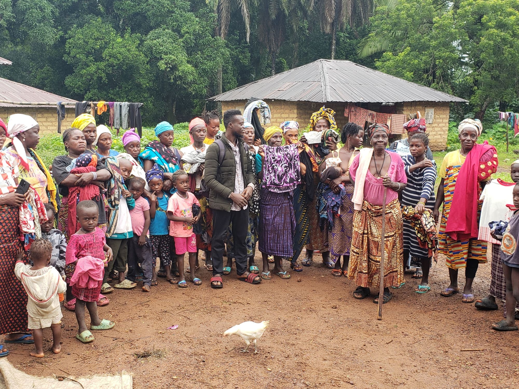 Women, men, and children from a village standing together in front of a mud-brick house surrounded by trees.