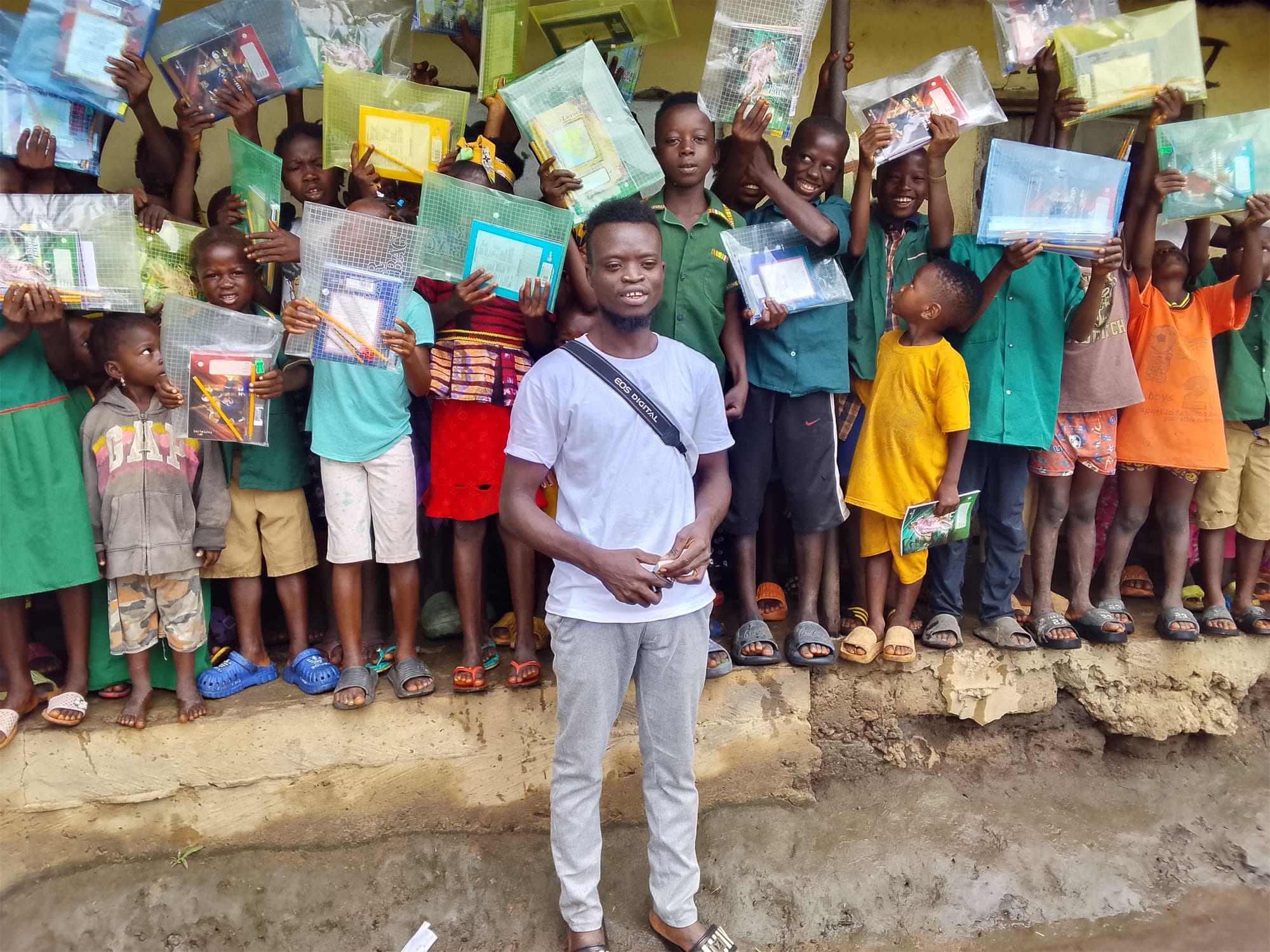 Children in school uniforms holding up clear pouches of notebooks and pencils around a man in a white T-shirt.